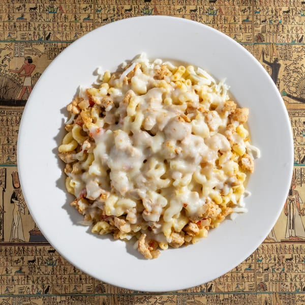 A white plate of cheesy macaroni and chicken sits on an Egyptian hieroglyphic-patterned tablecloth.