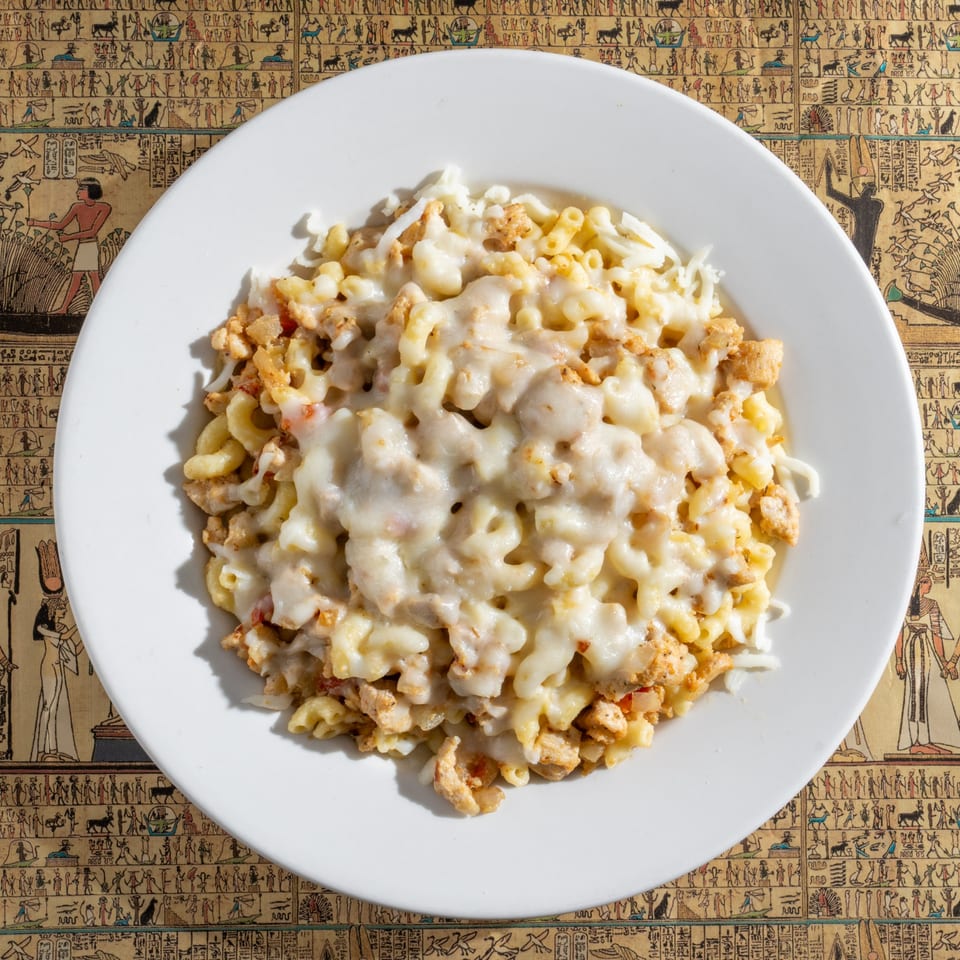 A white plate of cheesy macaroni and chicken sits on an Egyptian hieroglyphic-patterned tablecloth.