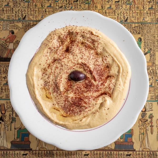 Creamy hummus with paprika and a black olive, served on a white plate over an Egyptian-themed tablecloth.