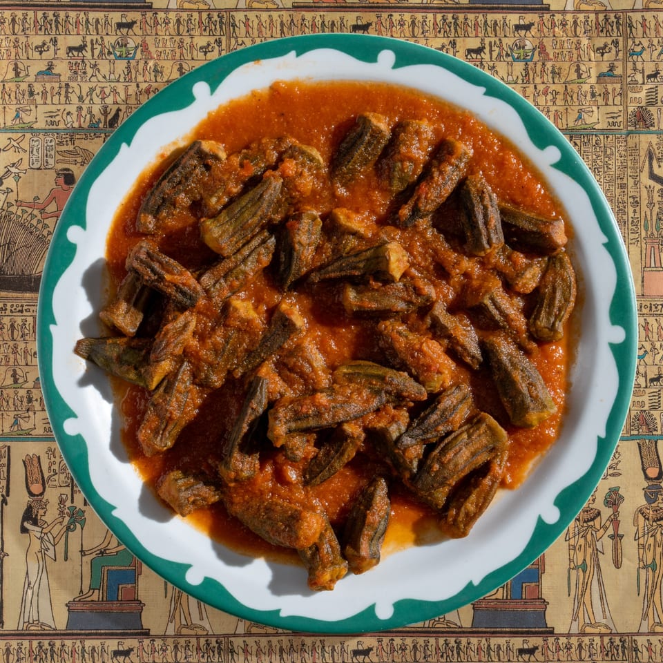 Egyptian okra stew in a white and green bowl, on a tablecloth with hieroglyphic patterns.