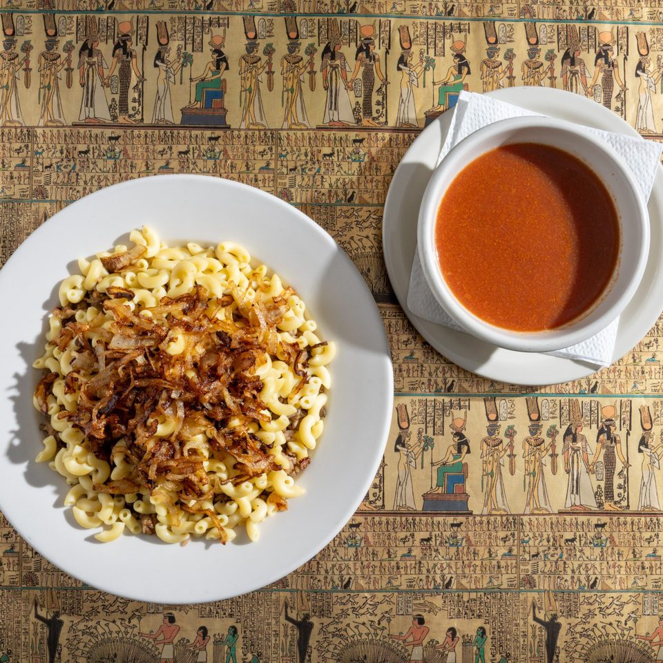 King Tut's Koshary, a pasta dish with crispy onions, and a side of tomato sauce on an Egyptian-themed tablecloth.